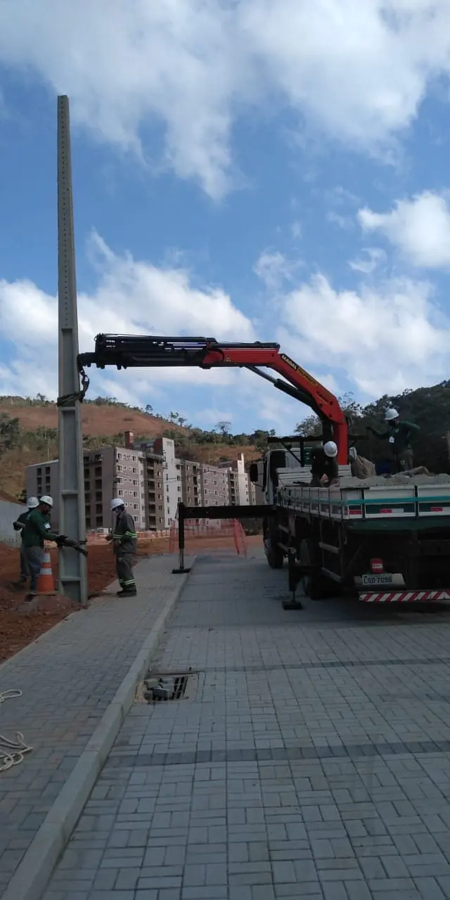 Poste de concreto sendo içado por guindauto, com equipe usando EPI